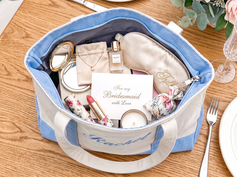 May include: A light blue and white canvas tote bag with the name Rachel embroidered on the front. The bag contains a gold compact mirror, a necklace in a pouch, a perfume bottle, a sleep mask, a floral lipstick, a candle, and a card reading For my Bridesmaid with Love.