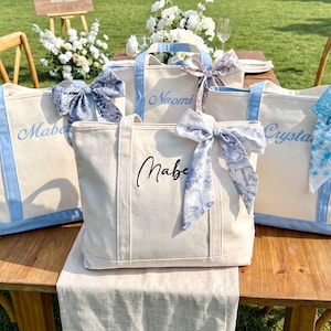 May include: Three off-white canvas tote bags with light blue trim and handles. Each bag features a decorative patterned ribbon tied to one handle and a personalized name embroidered or printed in script font on the front center of the bag.