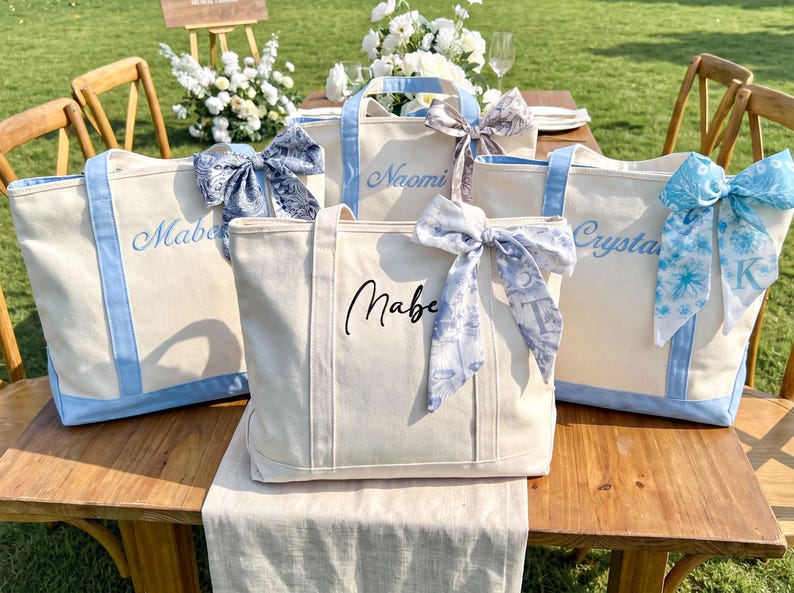 May include: Three off-white canvas tote bags with light blue trim and handles. Each bag features a decorative patterned ribbon tied to one handle and a personalized name embroidered or printed in script font on the front center of the bag.
