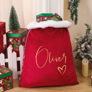 May include: A large red velvet Christmas sack with the name "Oliver" in gold script and a gold heart. The sack has a white faux fur trim. Several red and green Christmas-themed gift boxes and small Christmas trees are in the background.