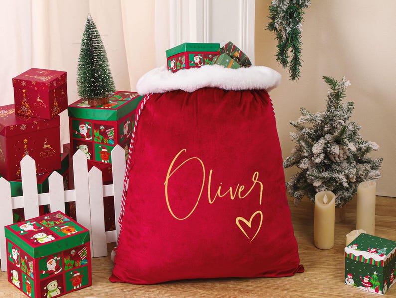 May include: A large red velvet Christmas sack with the name "Oliver" in gold script and a gold heart. The sack has a white faux fur trim. Several red and green Christmas-themed gift boxes and small Christmas trees are in the background.