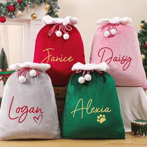 May include: Four plush Christmas gift sacks in red, pink, gray, and green. Each sack has a white trim and is personalized with a name in a cursive font. The green sack has a paw print. A Christmas tree and decorations are in the background.