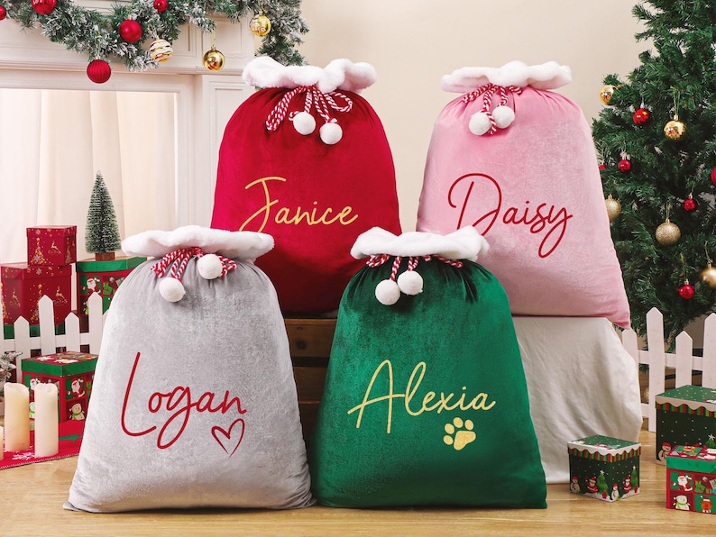 May include: Four plush Christmas gift sacks in red, pink, gray, and green. Each sack has a white trim and is personalized with a name in a cursive font. The green sack has a paw print. A Christmas tree and decorations are in the background.