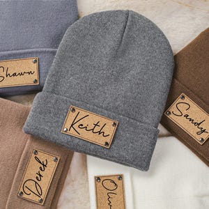 Könnte beinhalten: Fünf gestrickte Beanies in verschiedenen Farben mit personalisierten Kork-Namensschildern. Die Namen auf den Schildern sind Shawn, Keith, Sandy, Derek und Olin.