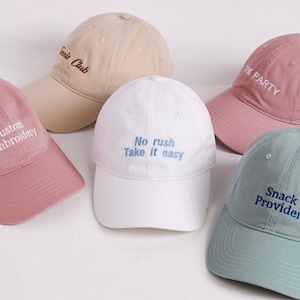 May include: Five baseball caps in various colours including beige, white, pink, and light blue. Each cap features embroidered text: 'Tennis Club', 'Custom Embroidery', 'No rush Take it easy', 'THE PARTY', and 'Snack Provider' in contrasting thread.