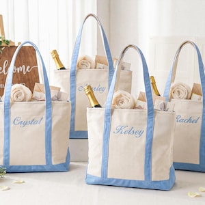 Peut inclure: Quatre sacs fourre-tout en toile blanc cassé avec des bordures et des anses bleu clair. Chaque sac porte un prénom brodé en écriture cursive bleu clair : Crystal, Marley, Kelsey et Rachel. Les sacs contiennent des serviettes blanches roulées.