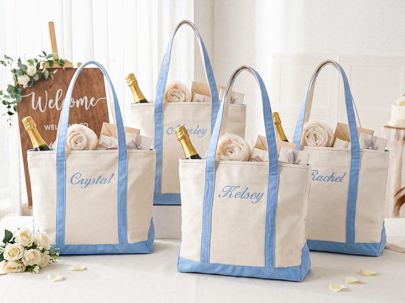 May include: Four off-white canvas tote bags with light blue trim and handles. Each bag features a name embroidered in light blue script: Crystal, Marley, Kelsey, and Rachel. The bags are filled with rolled white towels and champagne bottles.