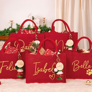 Puede incluir: Bolsas rojas con letras doradas de nombres como "Felix" e "Isabel". Cada bolsa tiene un llavero a juego con una etiqueta de madera y borlas decorativas. Las bolsas están llenas de regalos, perfectas para Navidad.