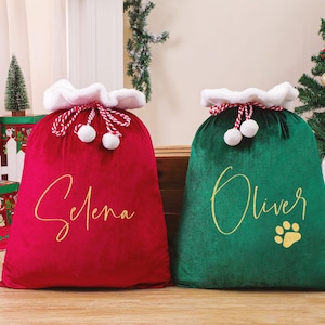 May include: Two plush Christmas gift bags, one red and one green. The red bag has the name "Selena" in gold script, and the green bag has "Oliver" and a paw print. Both bags have white faux fur trim and red and white drawstring closures.