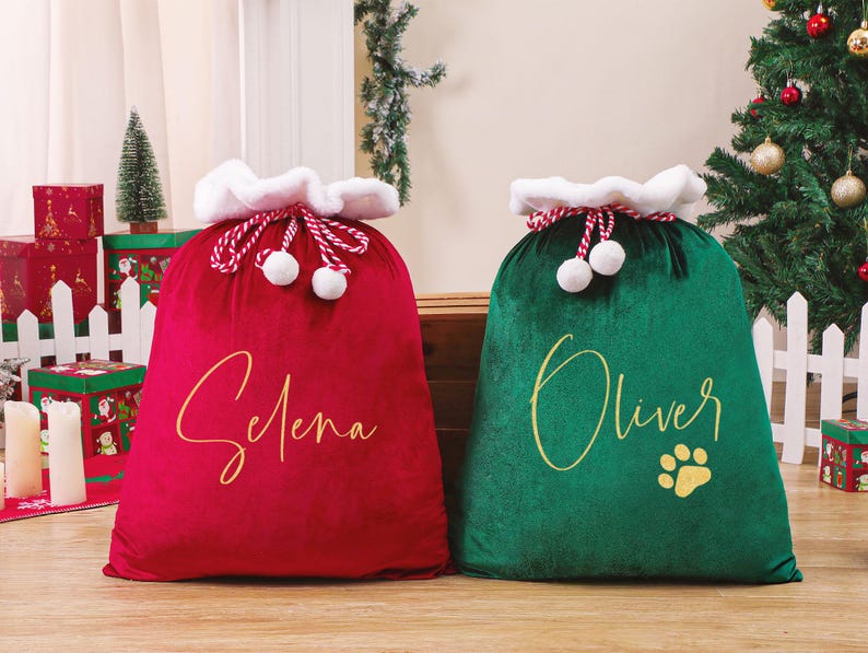 May include: Two plush Christmas gift bags, one red and one green. The red bag has the name "Selena" in gold script, and the green bag has "Oliver" and a paw print. Both bags have white faux fur trim and red and white drawstring closures.