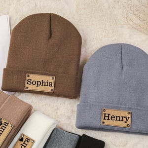 Op de afbeelding: Twee bruine en grijze gebreide mutsen met gepersonaliseerde naamstickers. De muts links heeft een sticker met de tekst "Sophia" en de muts rechts heeft een sticker met de tekst "Henry".