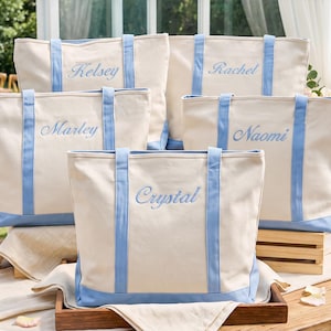 May include: Five off-white canvas tote bags with light blue trim and handles. Each bag features a unique name embroidered in light blue script: Kelsey, Rachel, Marley, Naomi, and Crystal. The bags are arranged in a group on a wooden surface.