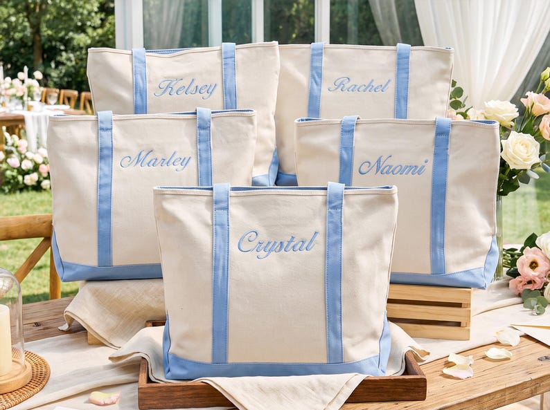 May include: Five off-white canvas tote bags with light blue trim and handles. Each bag features a unique name embroidered in light blue script: Kelsey, Rachel, Marley, Naomi, and Crystal. The bags are arranged in a group on a wooden surface.