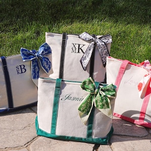 Puede incluir: Varias bolsas de lona personalizadas en crema, azul marino, verde y rosa. Cada bolsa presenta iniciales o un nombre y está adornada con un lazo decorativo.