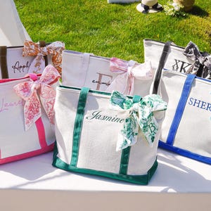 May include: White canvas tote bags with coloured trim and handles, personalised with names in various colours. Each bag is adorned with a decorative patterned bow. The bags are arranged on a white surface against a backdrop of green grass.
