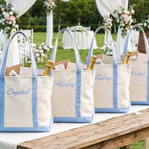 May include: Four off-white canvas tote bags with light blue trim and handles. Each bag features a name embroidered in light blue script: Crystal, Marley, Kelsey, and Rachel. Each bag contains a gold-colored bottle and various paper envelopes.