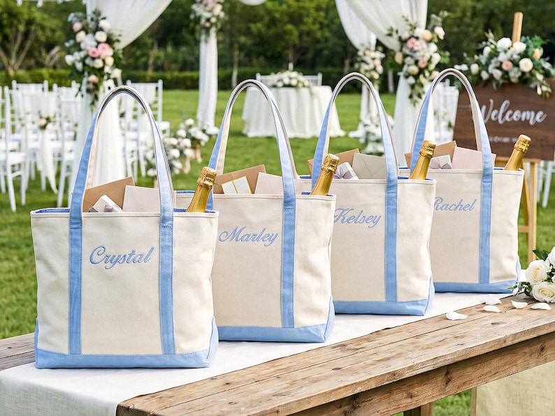 May include: Four off-white canvas tote bags with light blue trim and handles. Each bag features a name embroidered in light blue script: Crystal, Marley, Kelsey, and Rachel. Each bag contains a gold-colored bottle and various paper envelopes.
