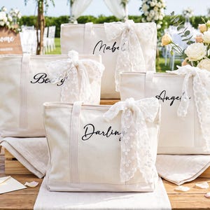 May include: Four off-white canvas tote bags with black cursive names printed on the front. Each bag features a white lace ribbon tied to the handle. The names printed on the bags are Mabel, Betty, Angela, and Darling. The bags have sturdy handles.