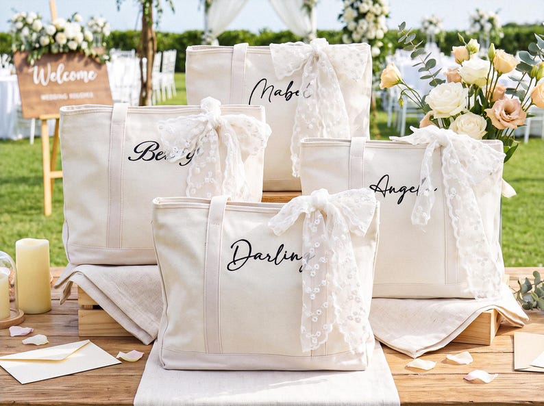May include: Four off-white canvas tote bags with black cursive names printed on the front. Each bag features a white lace ribbon tied to the handle. The names printed on the bags are Mabel, Betty, Angela, and Darling. The bags have sturdy handles.