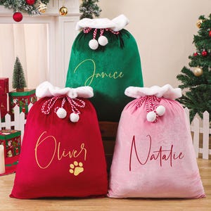 May include: Three plush Christmas gift sacks in red, green, and pink. Each sack has a white fluffy trim and a drawstring closure with white pom-poms. The red sack has the name "Oliver" and a paw print. The green sack has the name "Janice", and the pink sack has the name "Natalie".
