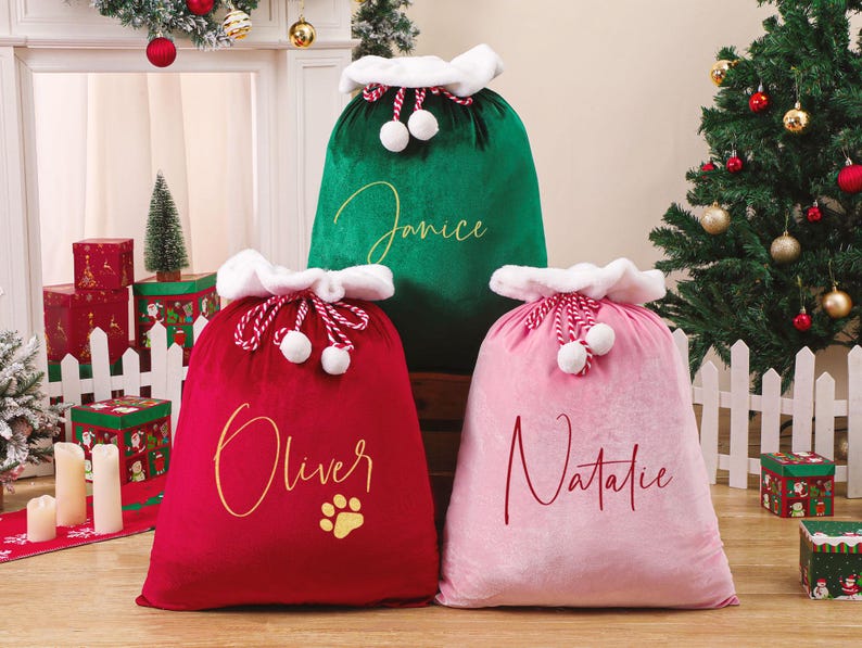 May include: Three plush Christmas gift sacks in red, green, and pink. Each sack has a white fluffy trim and a drawstring closure with white pom-poms. The red sack has the name "Oliver" and a paw print. The green sack has the name "Janice", and the pink sack has the name "Natalie".