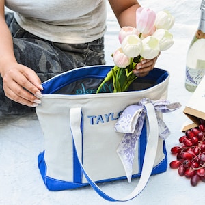May include: A canvas tote bag with blue accents and the name "TAYLOR" embroidered in blue. The bag is filled with flowers. A bottle of champagne and a paper bag with fruit are nearby.