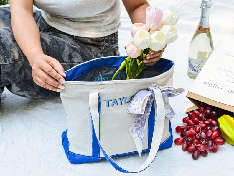 May include: A canvas tote bag with blue accents and the name "TAYLOR" embroidered in blue. The bag is filled with flowers. A bottle of champagne and a paper bag with fruit are nearby.