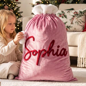 May include: A large pink velvet gift sack featuring a white faux fur trim at the top and red and white twisted rope drawstrings with white pom-poms. The name Sophia is embroidered in large, dark red cursive script on the front of the sack.