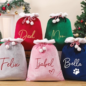 May include: Five plush Christmas gift bags in red, green, gray, pink, and blue. Each bag has a white trim and is personalized with a name or word in a contrasting color. The blue bag has a paw print.