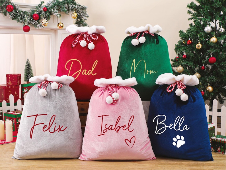 May include: Five plush Christmas gift bags in red, green, gray, pink, and blue. Each bag has a white trim and is personalized with a name or word in a contrasting color. The blue bag has a paw print.