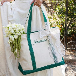 May include: A cream-colored canvas tote bag featuring dark green trim and handles. The name Jasmine is embroidered in dark green script on the front. A white lace ribbon is tied around one handle, and a bouquet of white calla lilies is inside.