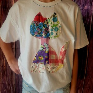K&ouml;nnte beinhalten: Wei&szlig;es T-Shirt mit einem bunten Patchwork-Katzen-Design. Die Katze besteht aus Stoffen mit Blumen-, Karo- und Mustermotiven in Rot, Blau, Lila und Rosa. Das T-Shirt hat einen Rundhalsausschnitt und kurze &Auml;rmel.