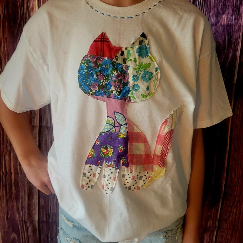 K&ouml;nnte beinhalten: Wei&szlig;es T-Shirt mit einem bunten Patchwork-Katzen-Design. Die Katze besteht aus Stoffen mit Blumen-, Karo- und Mustermotiven in Rot, Blau, Lila und Rosa. Das T-Shirt hat einen Rundhalsausschnitt und kurze &Auml;rmel.