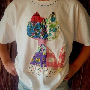 K&ouml;nnte beinhalten: Wei&szlig;es T-Shirt mit einem bunten Patchwork-Katzen-Design. Die Katze besteht aus verschiedenen gemusterten Stoffen in Rot-, Blau-, Lila- und Rosat&ouml;nen. Das Shirt hat einen Rundhalsausschnitt und kurze &Auml;rmel.