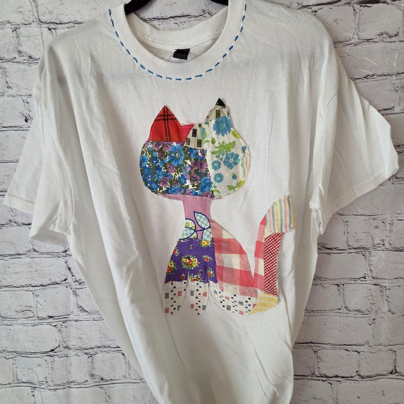 K&ouml;nnte beinhalten: Wei&szlig;es T-Shirt mit einem Patchwork-Katzen-Design. Die Katze besteht aus verschiedenen gemusterten Stoffen in Rot, Blau, Lila und Rosa. Der Halsausschnitt hat eine blaue Naht. Das Shirt h&auml;ngt an einem Kleiderb&uuml;gel vor einer Backsteinmauer.