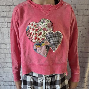 May include: A pink sweatshirt with a distressed look, featuring four heart-shaped patches in various patterns. The patches include floral, gingham, and patchwork designs. The sweatshirt has a crew neck and long sleeves.