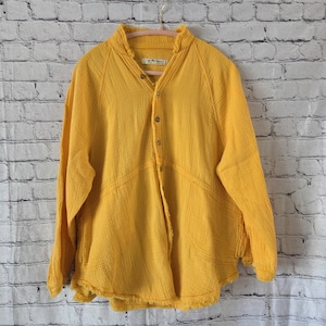 Pu&ograve; includere: Camicia senape a maniche lunghe con abbottonatura e colletto alla coreana. La camicia ha un tessuto strutturato e dettagli di cucitura decorativi. La camicia &egrave; appesa a una gruccia rosa contro un muro di mattoni bianchi.
