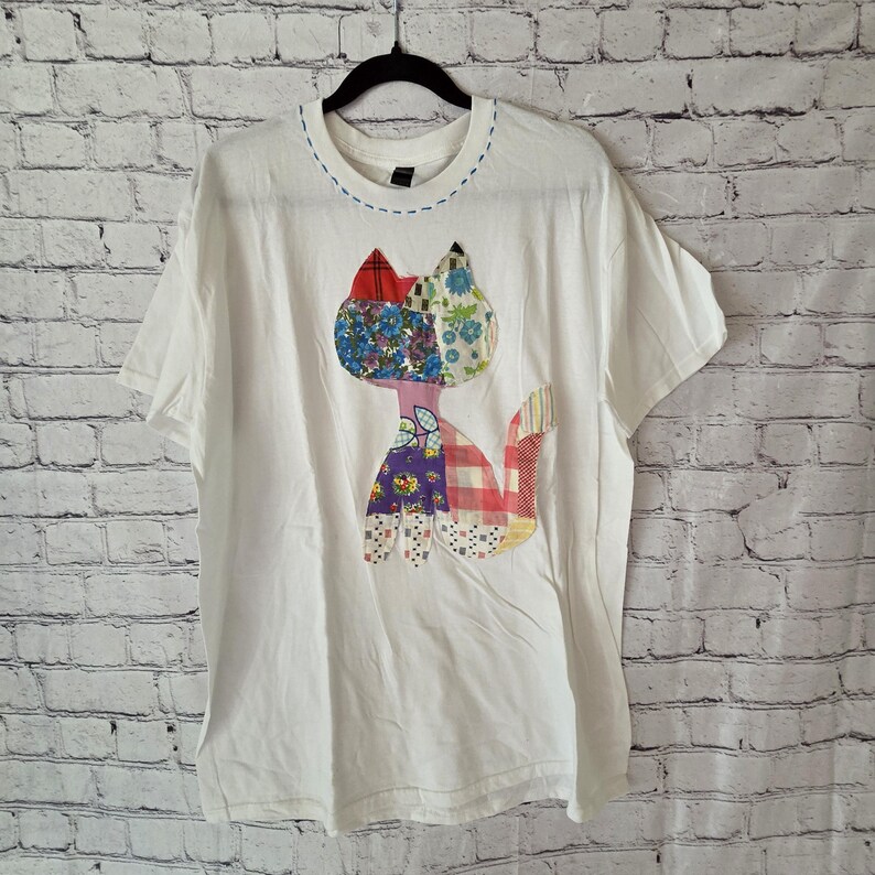 K&ouml;nnte beinhalten: Wei&szlig;es T-Shirt mit einem Patchwork-Katzen-Design. Die Katze hat eine Vielzahl von bunten Stoffmustern, darunter Blumen-, Karo- und Unifarben. Der Halsausschnitt hat eine blaue Naht.