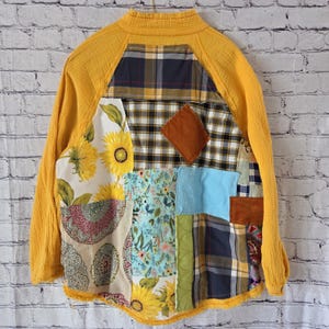 Pu&ograve; includere: Giacca patchwork con colletto e maniche gialle. La giacca presenta una variet&agrave; di motivi di tessuto, tra cui girasoli, stampe floreali, plaid e una toppa a forma di diamante marrone. La giacca &egrave; appesa a un muro di mattoni bianchi.