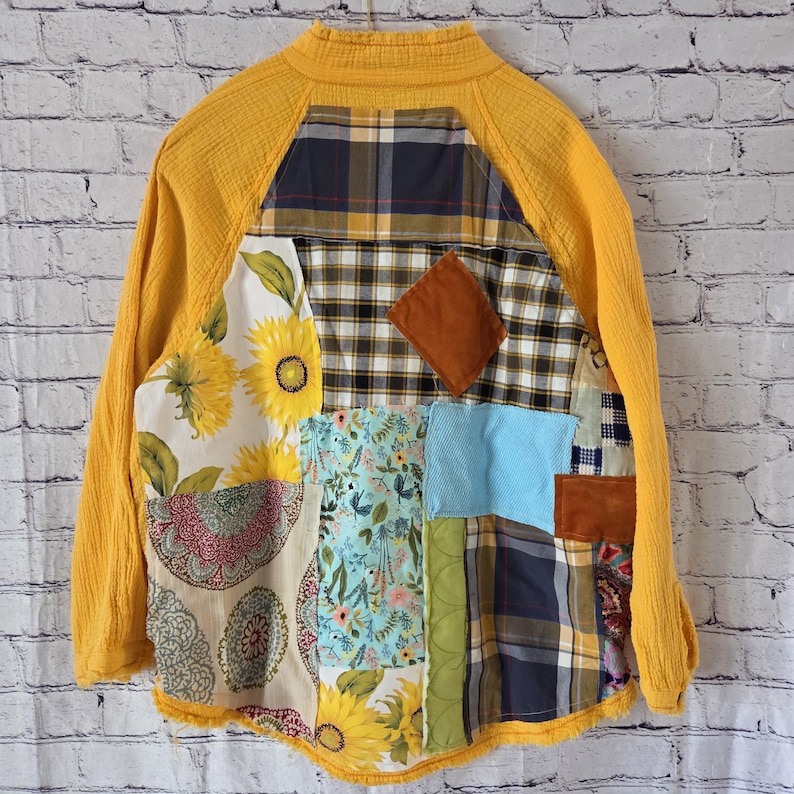 Pu&ograve; includere: Giacca patchwork con colletto e maniche gialle. La giacca presenta una variet&agrave; di motivi di tessuto, tra cui girasoli, stampe floreali, plaid e una toppa a forma di diamante marrone. La giacca &egrave; appesa a un muro di mattoni bianchi.