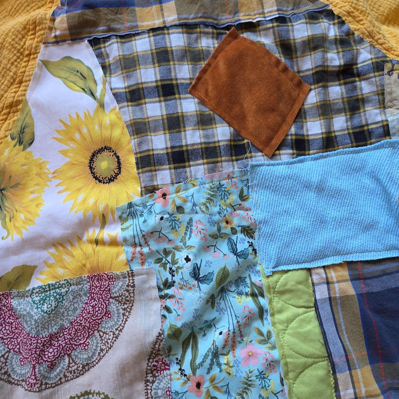 Pu&ograve; includere: Una trapunta patchwork con una variet&agrave; di tessuti. La trapunta presenta una stampa di girasoli, motivi floreali, quadri e colori uniti. Un quadrato di tessuto marrone &egrave; cucito sulla trapunta. La trapunta &egrave; un mix di trame e colori.