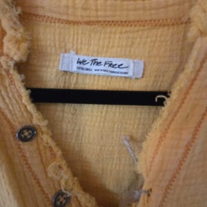 Pu&ograve; includere: Primo piano di una camicia gialla con bottoni e tessuto strutturato. La camicia ha un design con bordi sfilacciati e bottoni decorativi. L'etichetta recita "We The Free". La camicia &egrave; appesa a una gruccia nera.