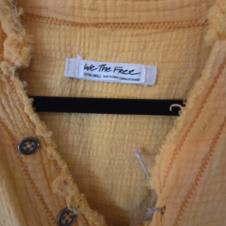 Pu&ograve; includere: Primo piano di una camicia gialla con bottoni e tessuto strutturato. La camicia ha un design con bordi sfilacciati e bottoni decorativi. L'etichetta recita "We The Free". La camicia &egrave; appesa a una gruccia nera.