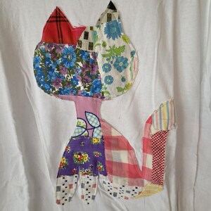 K&ouml;nnte beinhalten: Wei&szlig;es T-Shirt mit einem Patchwork-Katzen-Design. Die Katze besteht aus verschiedenen Stoffmustern, darunter Blumen-, Karo- und geometrische Drucke in Rot-, Blau-, Lila- und Rosat&ouml;nen. Der Schwanz der Katze ist ausgestreckt, und das Design ist auf dem Shirt zentriert.