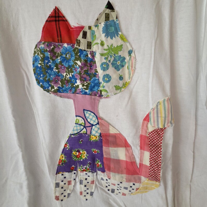 K&ouml;nnte beinhalten: Wei&szlig;es T-Shirt mit einem Patchwork-Katzen-Design. Die Katze besteht aus verschiedenen Stoffmustern, darunter Blumen-, Karo- und geometrische Drucke in Rot-, Blau-, Lila- und Rosat&ouml;nen. Der Schwanz der Katze ist ausgestreckt, und das Design ist auf dem Shirt zentriert.
