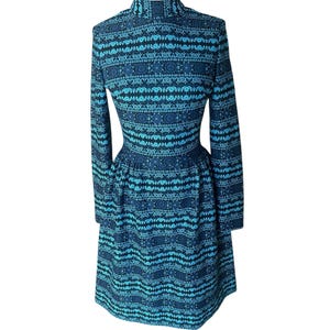 May include: A long-sleeved, knee-length dress with a high neckline. The dress features a repeating pattern of turquoise and navy blue shapes. The dress is displayed on a white mannequin, showcasing its fitted bodice and flared skirt.