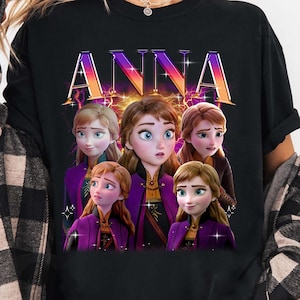 Puede incluir: Camiseta negra con la palabra "ANNA" en una fuente colorida y estilizada. La camiseta muestra un gr&aacute;fico de cinco personajes animados con piel clara, vestidos con trajes morados y dorados. Los personajes tienen diferentes expresiones y peinados.