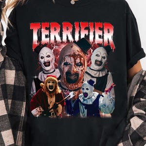 May include: Black t-shirt featuring the word "TERRIFIER" in red, above a graphic of several scary clowns. The clowns have white faces, black and white outfits, and menacing expressions. The shirt is a casual style.