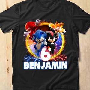 Könnte beinhalten: Schwarzes T-Shirt mit einer farbenfrohen Grafik von Sonic the Hedgehog-Charakteren. Das Design enthält die Zahl "6" und den Namen "BENJAMIN" in Weiß, umgeben von einem goldenen und blauen Kreis mit Blitzen.