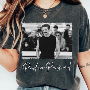 May include: Dark gray t-shirt featuring a black and white image of a man with the text "Pedro Pascal" in a cursive font. The image is framed with a white border. The shirt is a casual style.